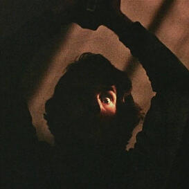 A silhouette of a man with shoulder-length hair holding something above his head with only one wide eye visible