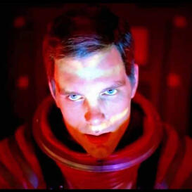 A man with short brown hair in a red spacesuit with harsh red lighting with his head tilted down, making a serious or stern expression at the camera