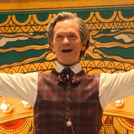 A man with pushed-back gray hair wearing a white dress shirt and reddish-brown vest with his arms outstretched