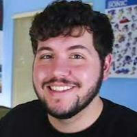 A man with curly black hair and light facial hair wearing a black t-shirt and smiling at the camera